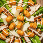 An overhead view of a large bowl of crispy tater caesar salad with golden smashed tater tot croutons, chicken, and parmesan cheese.